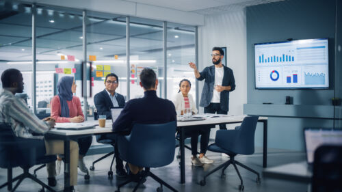 Diverse Modern Office: Businessman Leads Business Meeting with Managers, Talks, uses Presentation TV with Statistics, Infographics. Digital Entrepreneurs Work on e-Commerce Project.
