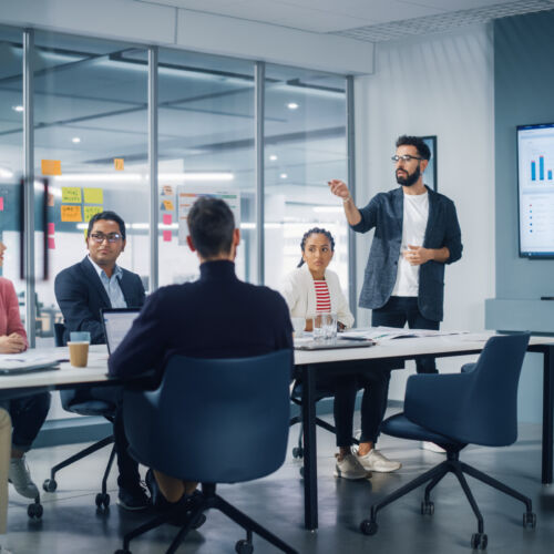 Diverse Modern Office: Businessman Leads Business Meeting with Managers, Talks, uses Presentation TV with Statistics, Infographics. Digital Entrepreneurs Work on e-Commerce Project.